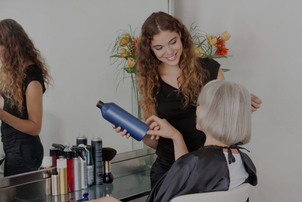 Eine lächelnde junge Friseurin mit lockigem Haar hält eine blaue Haarpflegeflasche und berät eine ältere Frau mit silbergrauem Haar, die auf einem Stuhl im Salon sitzt.