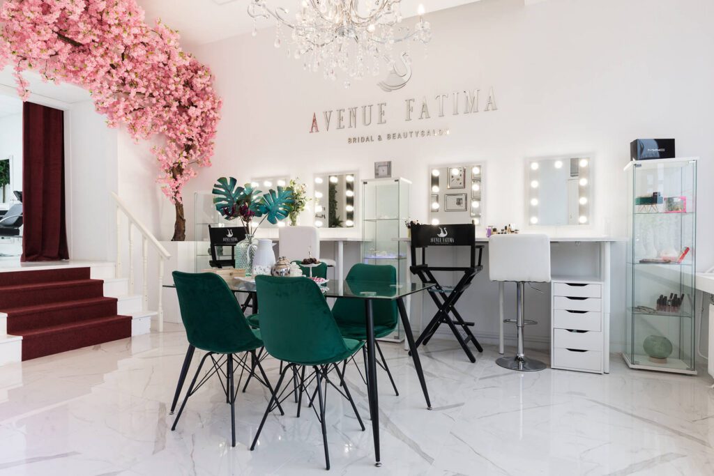 Ein heller, eleganter Braut- und Schönheitssalon namens „Avenue Fatima“. Der Raum verfügt über mehrere Schminkplätze mit beleuchteten Spiegeln, smaragdgrüne Samtstühle um einen Glastisch und einen dekorativen rosa Kirschblütenbaum.
